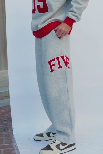 mens, Close-up of a person wearing light heather gray sweatpants with red trim and the word "FIVE" in red varsity-style lettering on the leg, holding up a matching heather gray sweatshirt with red trim and a large red number.