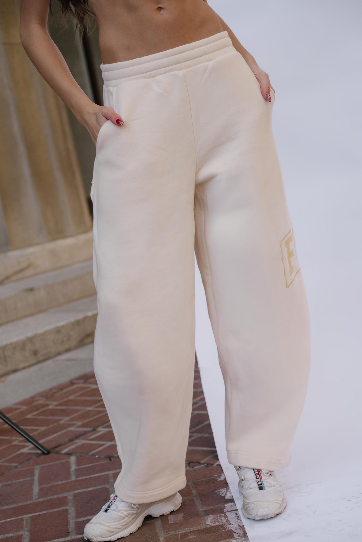 kids, A girl wearing loose, wide-leg butter yellow sweatpants with side pockets and a large yellow-toned logo embroidered on the lower left leg, paired with white sneakers, standing outdoors on a brick and concrete surface.