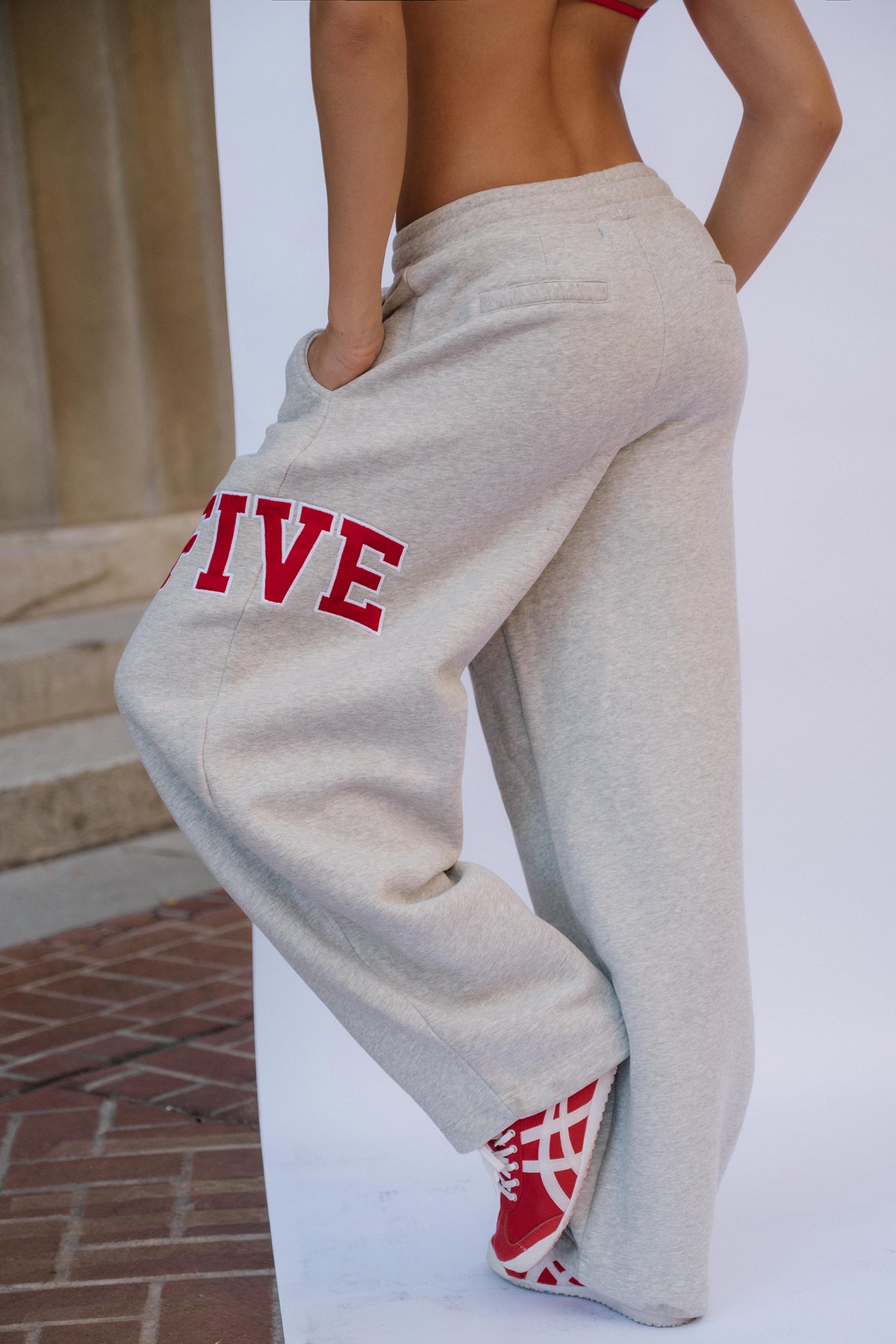 teen, A teen girl wearing light heather gray sweatpants with red collegiate-style lettering on the leg, shown from the back in an outdoor setting with a white wall and brick steps in the background.