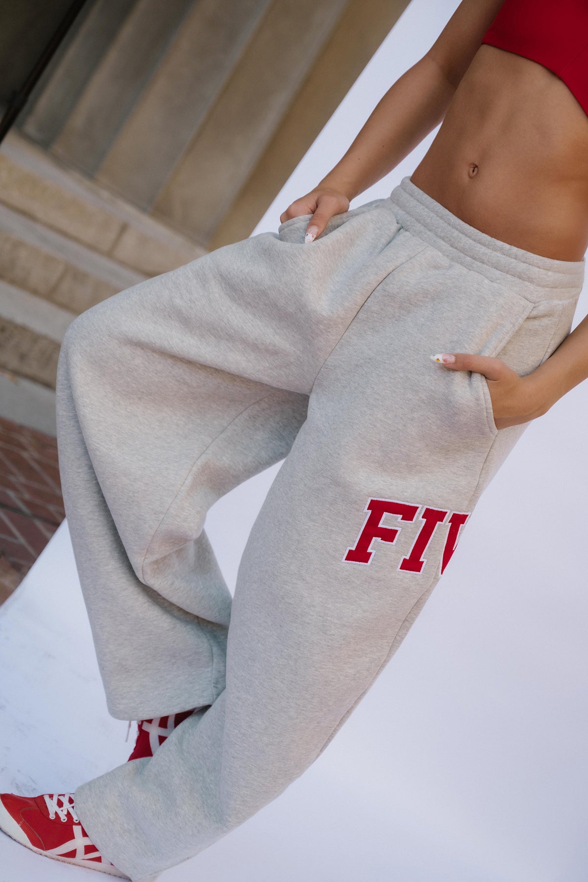 teen, A girl wearing light heather gray sweatpants with red collegiate-style lettering on the lower leg, paired with red sneakers, posed against a white wall.