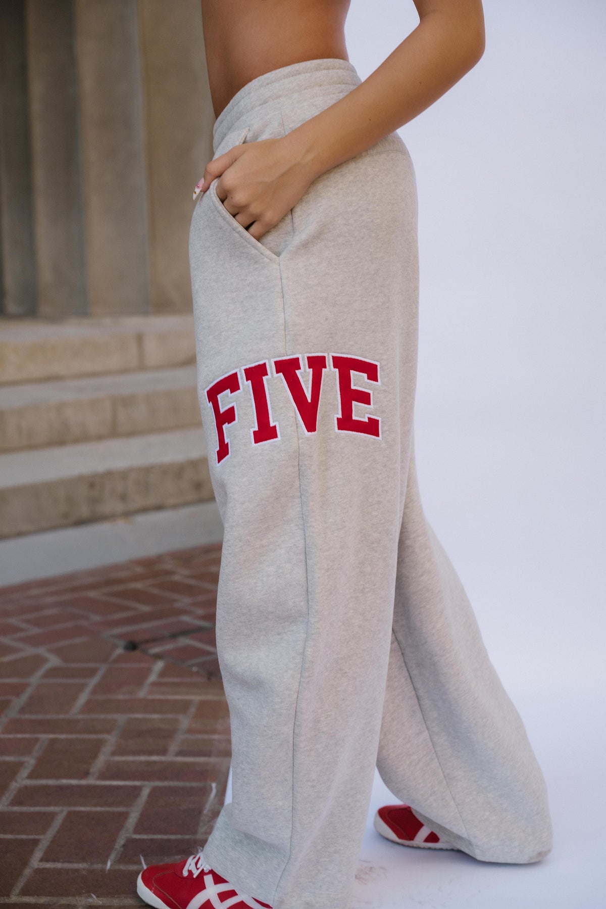 teen, A girl wearing light heather gray sweatpants with the word 'FIVE' in red collegiate block letters outlined in white on the left leg, standing outdoors near stone steps and a brick floor.