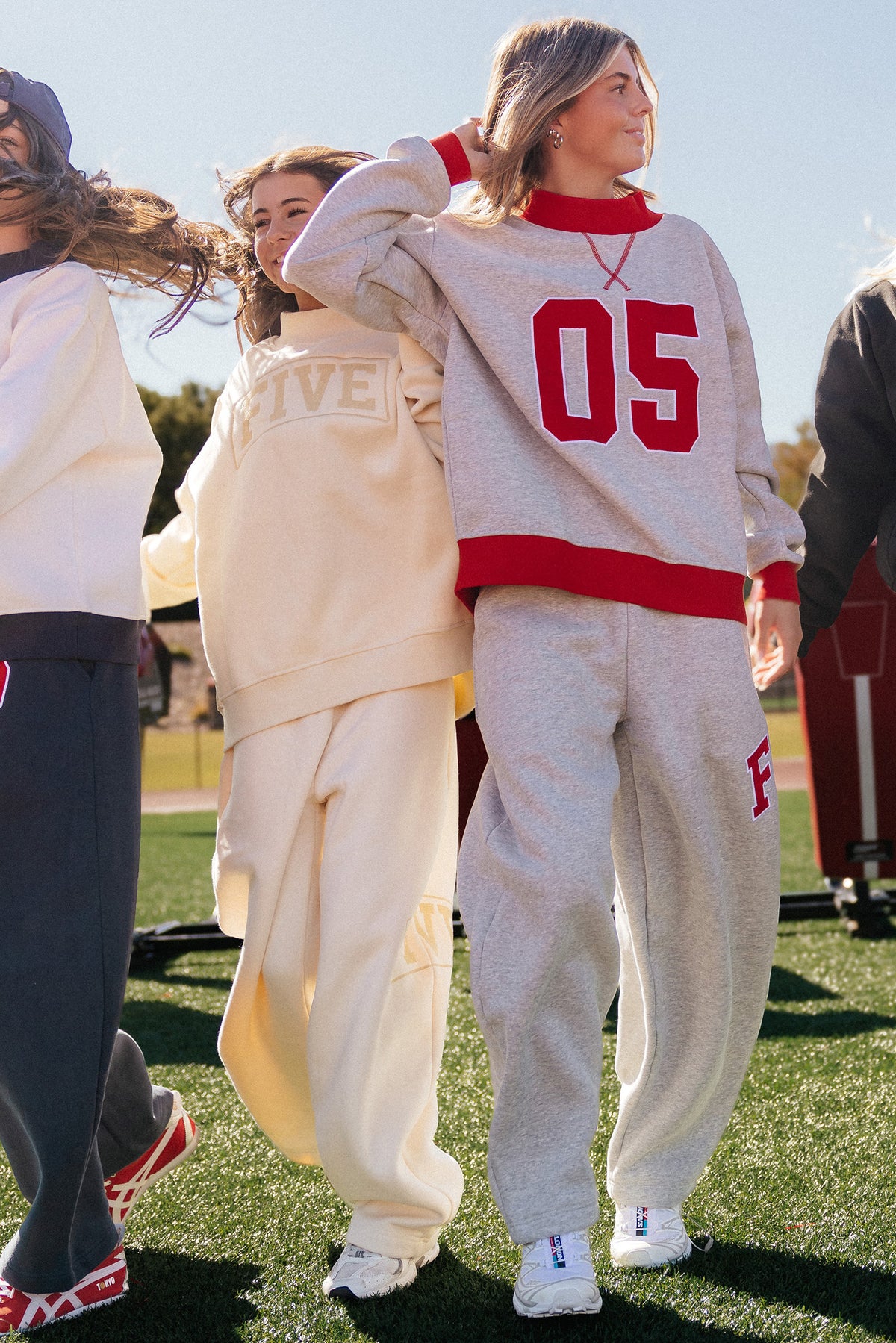 teen, Two girls walking outdoors on grass wearing matching sweatshirt and sweatpants sets. The girl in front wears a light heather gray set with bold red trim and large red '05' lettering on the sweatshirt, while the girl behind wears a cream set with 'FIVE' on the chest.
