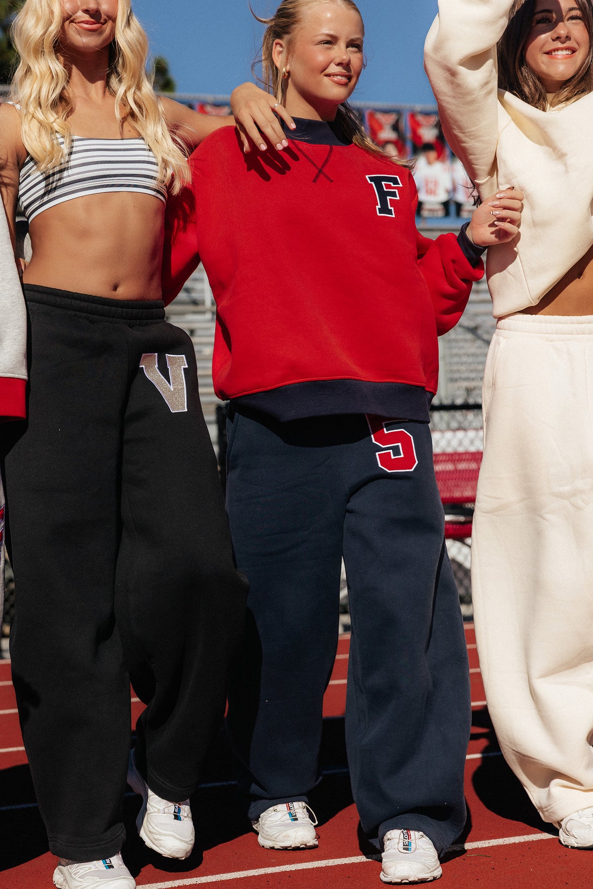 teen, Three women standing together on a red running track at a stadium, wearing matching athletic sweatpants in navy, , and yellow, paired with red, white, and striped crop tops. The center woman wears a red sweatshirt with a large letter 'F' on the chest and navy sweatpants with a red letter 'S'.