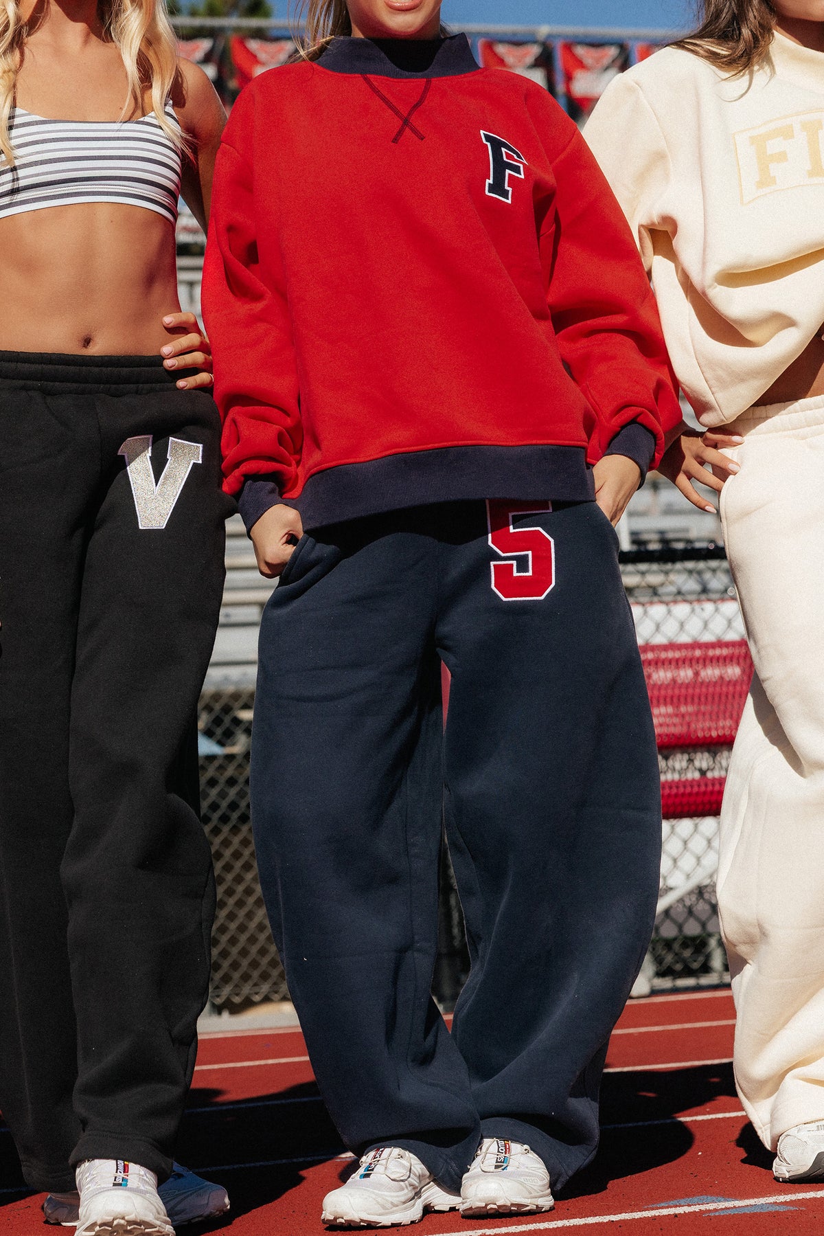 teen, standing outdoors on a track wearing an oversized red sweatshirt with a navy collar and hem, navy sweatpants with a red number 5 on the thigh, and white sneakers. Other people are partially visible at the edges.