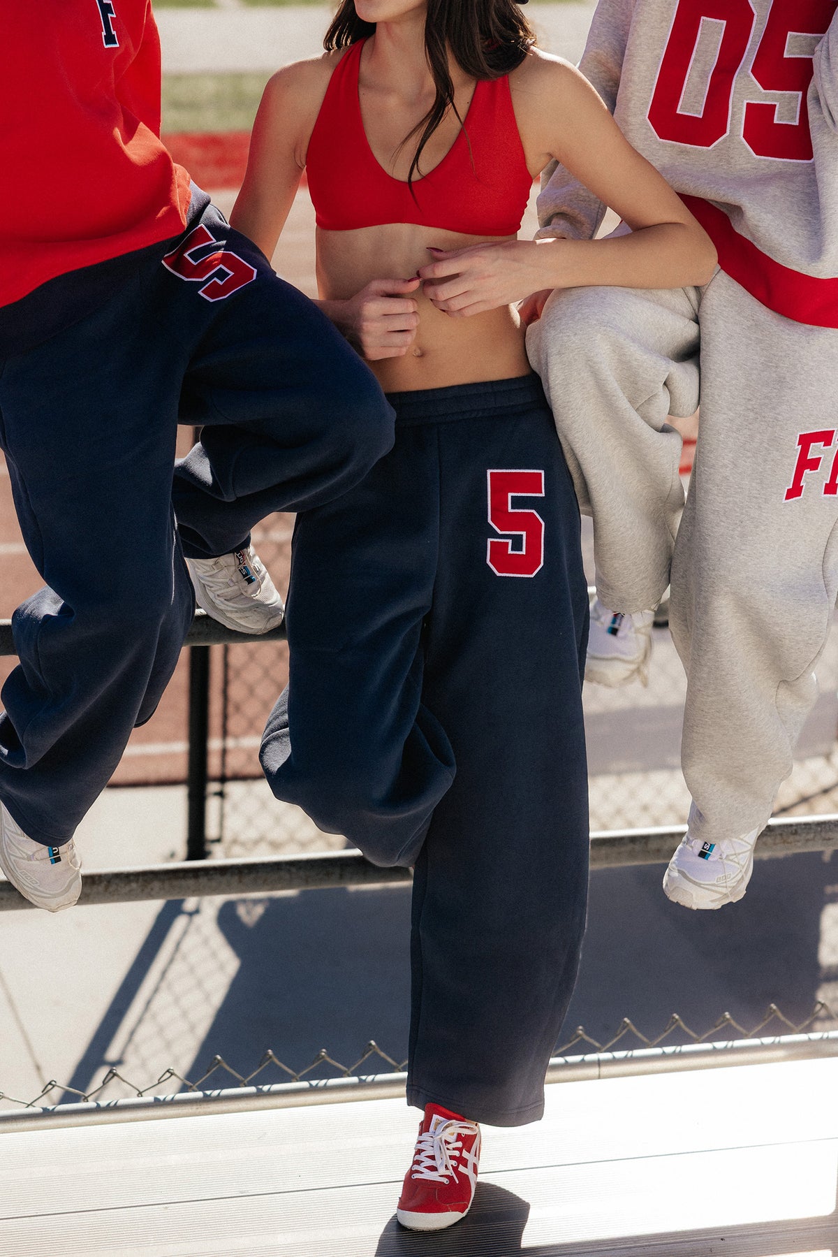 kids, A girl outdoors wearing loose navy sweatpants with a red number 5 on the thigh, a red sports bra, and red sneakers, with other people partially visible in athletic clothing around them.