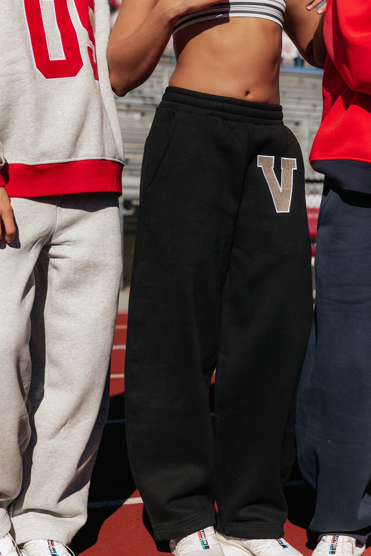 teen, Close-up of a girl wearing black sweatpants with a large metallic silver letter 'V' patch on the left thigh, standing on an outdoor track or field. Parts of two other people are visible at the left and right edges.