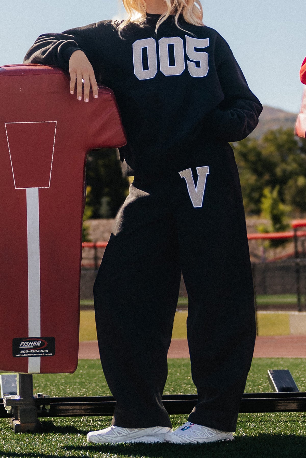 teen, A girl outdoors on a sports field wearing a black sweatshirt and matching sweatpants with large metallic silver varsity-style lettering, leaning against a red football pad.