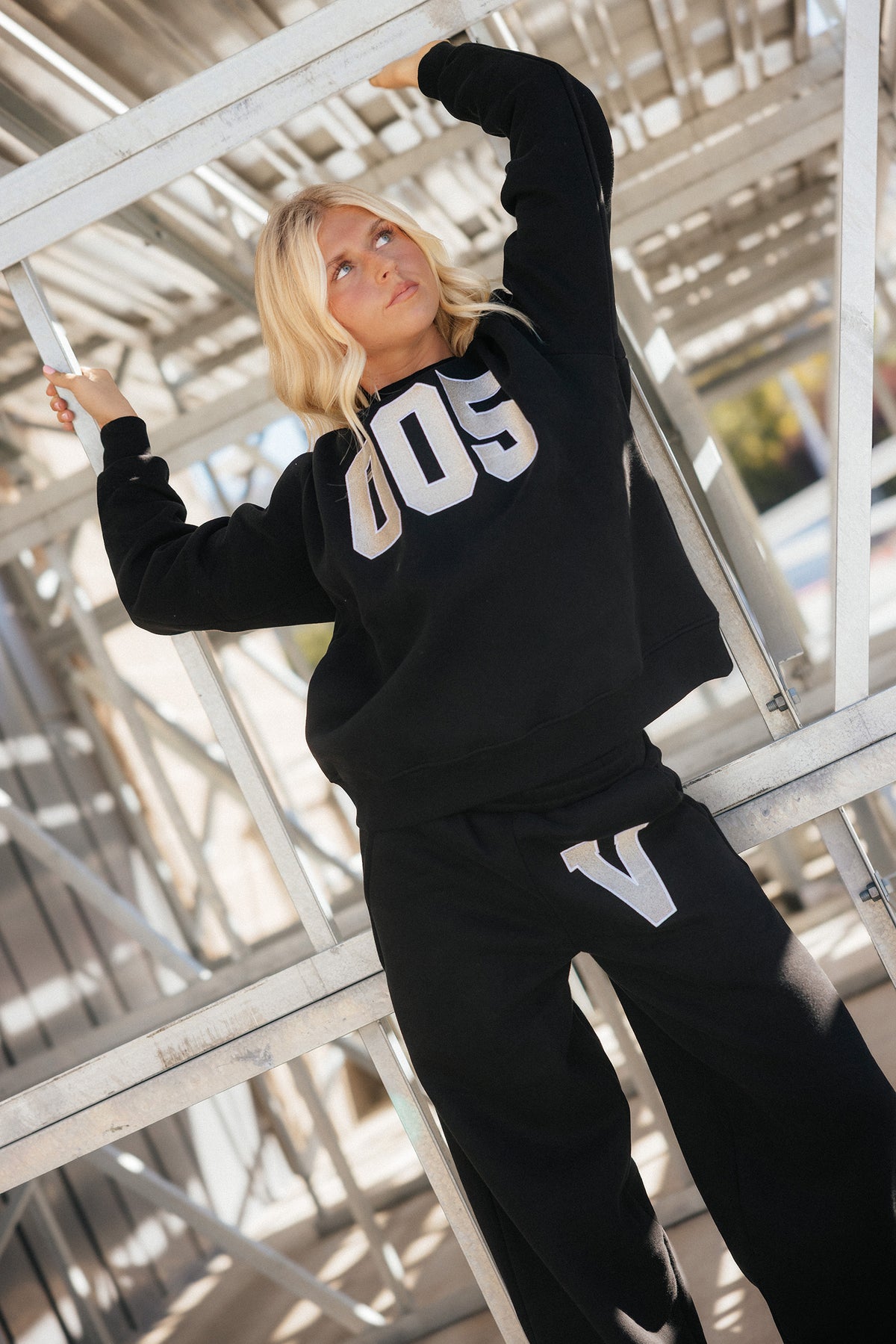 teen, A blonde girl wearing a black sweatshirt and matching sweatpants with metallic silver varsity-style lettering, posing on metal scaffolding.