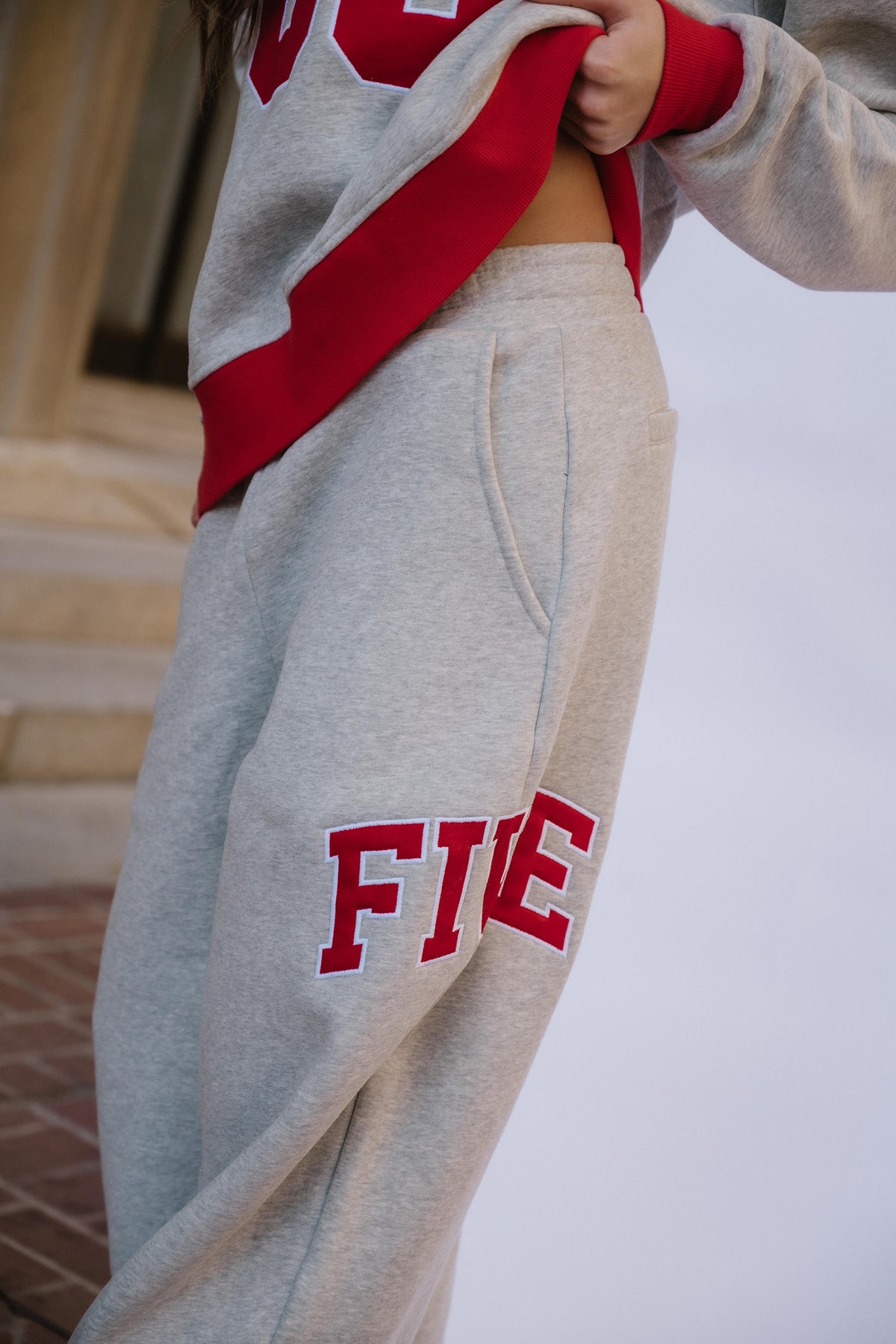 kids, A girl wearing a light heather gray sweatshirt and matching sweatpants with bold red collegiate lettering on the pants reading 'FIVE'. The photo is cropped to the torso and legs, with a blurred outdoor architectural background.