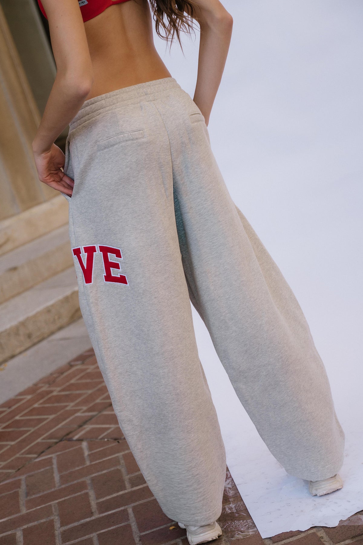 kids, A girl wearing light heather gray wide-leg sweatpants with red embroidered letters 'FIVE' on the left leg, photographed outdoors against a white backdrop with brick flooring visible.