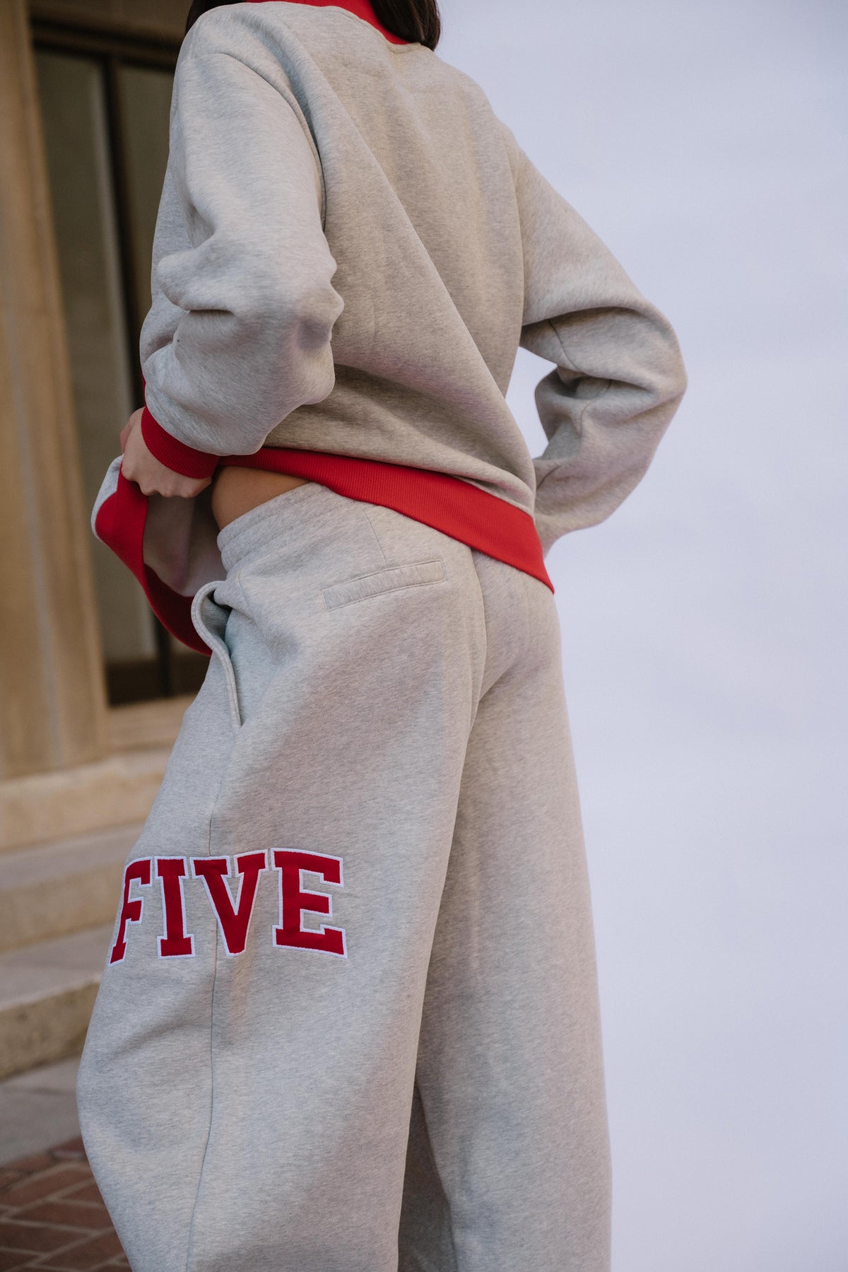 kids, A girl shown from the back wearing a light gray sweatshirt and matching sweatpants with bold red trim and red lettering on the leg, photographed outdoors against a pale sky and building facade.
