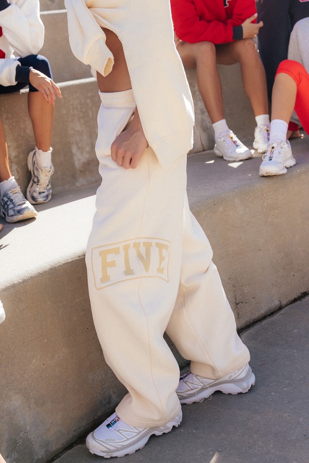 teen, A girl wearing butter yellow  sweatpants with a large beige 'FIVE' graphic embroidered on the left leg, styled with white sneakers, standing outdoors on concrete steps with other people visible in the background.