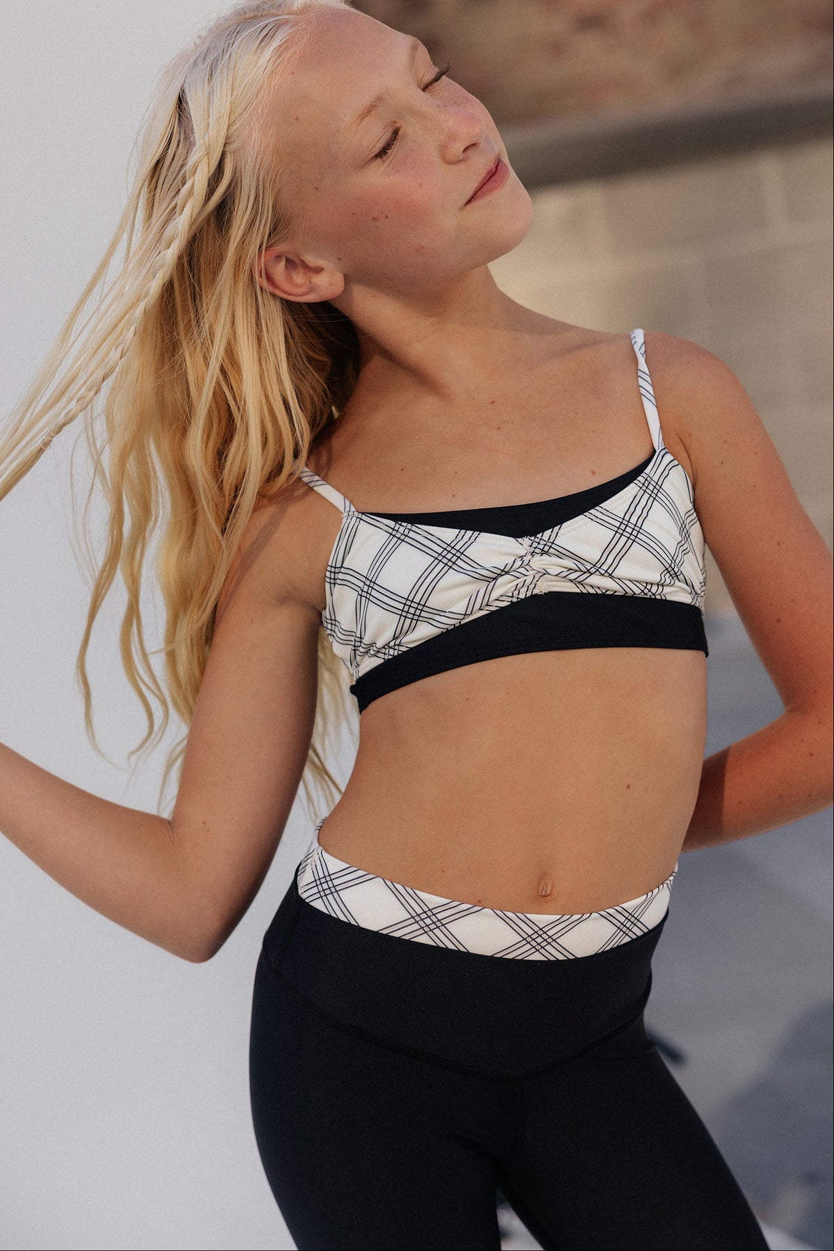 kids, A young blonde girl wearing a black and white plaid sports bra and matching black leggings, posing outdoors against a light gray wall.